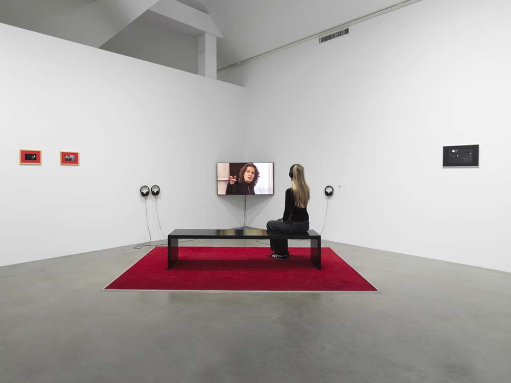 red carpet, TV screen, speakers, picture frames, person watching video on bench.